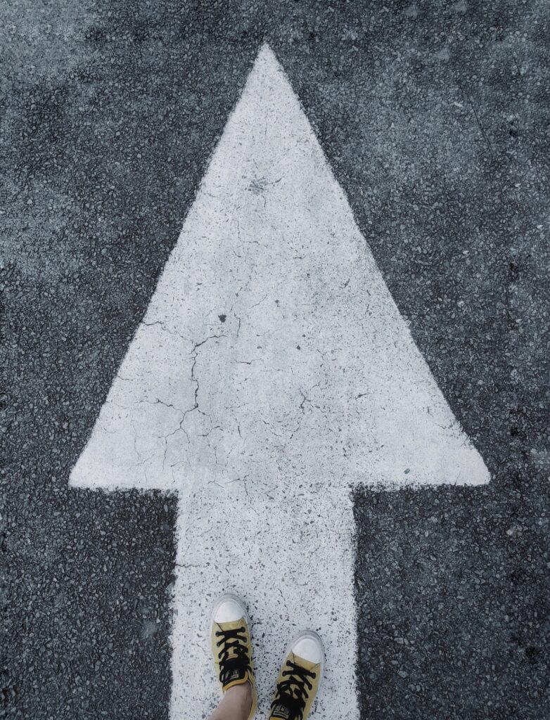 pexels-photo-1745766-1745766 Top-down view of white arrow on asphalt with sneakers stepping forward.